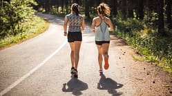 Laufen im Sommer: Zwei junge Frauen joggen auf einer sonnigen Straße durch einen Wald.