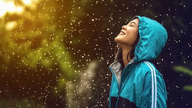 Laufmotivation bei jedem Wetter: Junge Läuferin steht mit einer Regenjacke glücklich im Regen.