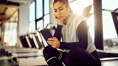 Laufdruck durch Social Media: Eine Läuferin sitzt im Fitnessstudio auf einem Laufband und schaut auf ihr Smartphone.
