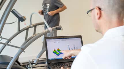 Ein Mann blickt auf einen Computerbildschirm, auf dem Daten zu sehen sind. Im Hintergrund läuft eine weitere Person auf einem Laufband. Der Mann am PC deutet auf den Bildschirm. Ein Mann blickt auf einen Computerbildschirm, auf dem Daten zu sehen sind. Im Hintergrund läuft eine weitere Person auf einem Laufband. Der Mann am PC deutet auf den Bildschirm.