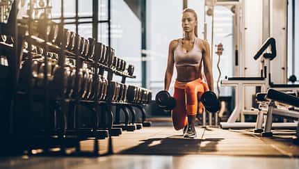 Female athlete exercising with dumbbells in a lunge position at gym.