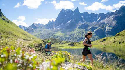 Zwei Teilnehmende laufen bei sonnigem Wetter auf einem Marathon-Trail entlang eines Bergsees. Im Hintergrund ist das Gebirgspanorama des Montafon zu erkennen.
