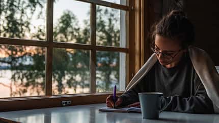Abendroutine: Eine Frau sitzt mit einer Tasse Tee am Tisch und schreibt in ein Tagebuch.