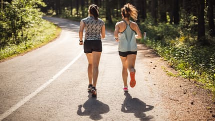Laufen im Sommer: Zwei junge Frauen joggen auf einer sonnigen Straße durch einen Wald.