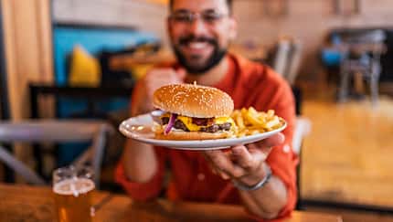 Häufige Ernährungsfehler nach dem Training: Ein sportlicher Mann bekommt einen Teller mit einem Burger und Pommes, auf dem Tisch steht ein Glas Bier.