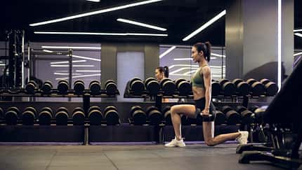 Metcon-Training: Kraft trifft Ausdauer. Eine Frau trainiert im Fitnessstudio Ausfallschritte mit Hanteln.