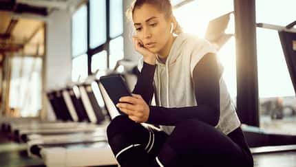 Laufdruck durch Social Media: Eine Läuferin sitzt im Fitnessstudio auf einem Laufband und schaut auf ihr Smartphone.