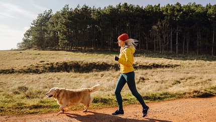 Eine Läuferin joggt entspannt mit ihrem Hund durch eine schöne Landschaft mit Feldern und Wald. Eine Läuferin joggt entspannt mit ihrem Hund durch eine schöne Landschaft mit Feldern und Wald.
