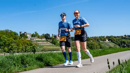 Eine Läuferin und ein Läufer mit grüner Landschaft und blauem Himmel im Hintergrund. Eine Läuferin und ein Läufer mit grüner Landschaft und blauem Himmel im Hintergrund.