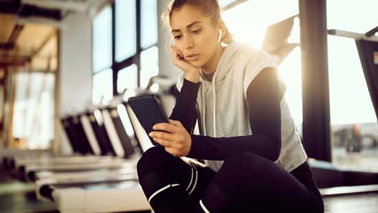 Laufdruck durch Social Media: Eine Läuferin sitzt im Fitnessstudio auf einem Laufband und schaut auf ihr Smartphone.