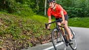 Eine junge Frau fährt Rennrad auf einer Straße durch den Wald. Triathlon-Trainingsplan: Olympische Distanz schaffen.