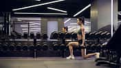 Metcon-Training: Kraft trifft Ausdauer. Eine Frau trainiert im Fitnessstudio Ausfallschritte mit Hanteln.