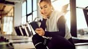 Laufdruck durch Social Media: Eine Läuferin sitzt im Fitnessstudio auf einem Laufband und schaut auf ihr Smartphone.