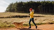 Eine Läuferin joggt entspannt mit ihrem Hund durch eine schöne Landschaft mit Feldern und Wald.